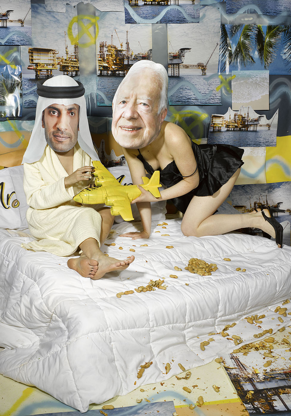 Two figures wearing masks of US former president Jimmy Carter and UAE oil minister in bed together holding a yellow model airplane. Discarded peanut shells litter the scene with a collage backdrop of oil rigs in water.