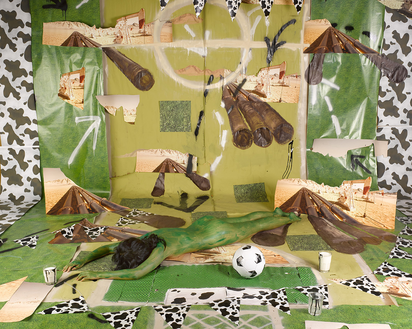 Collage; a nude figure painted green laying on a scene with oil pipelines, a football field, and a jersey cow pattern.