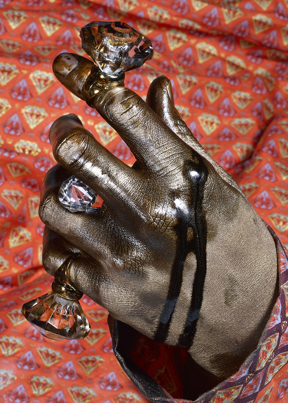 Closeup of a hand covered in gold paint and black oil, wearing two large diamond rings and holding an additional gemstone between the middle fingers.