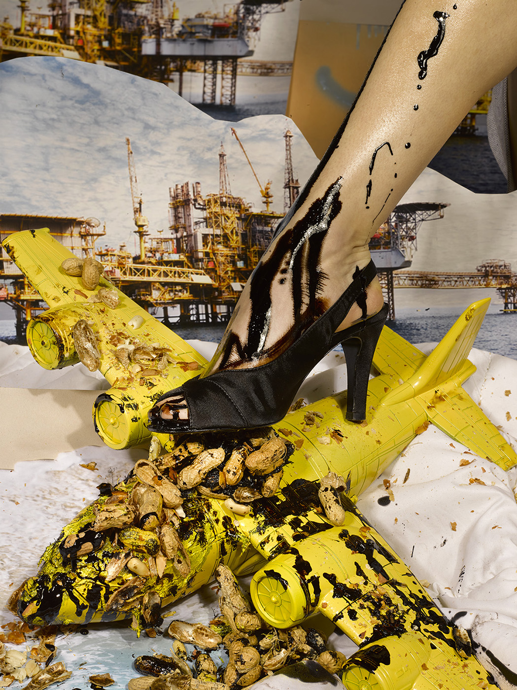 Closeup of a black stiletto stepping on a yellow model plane covered in oil and peanut shells, with an oil rig collage in the background.