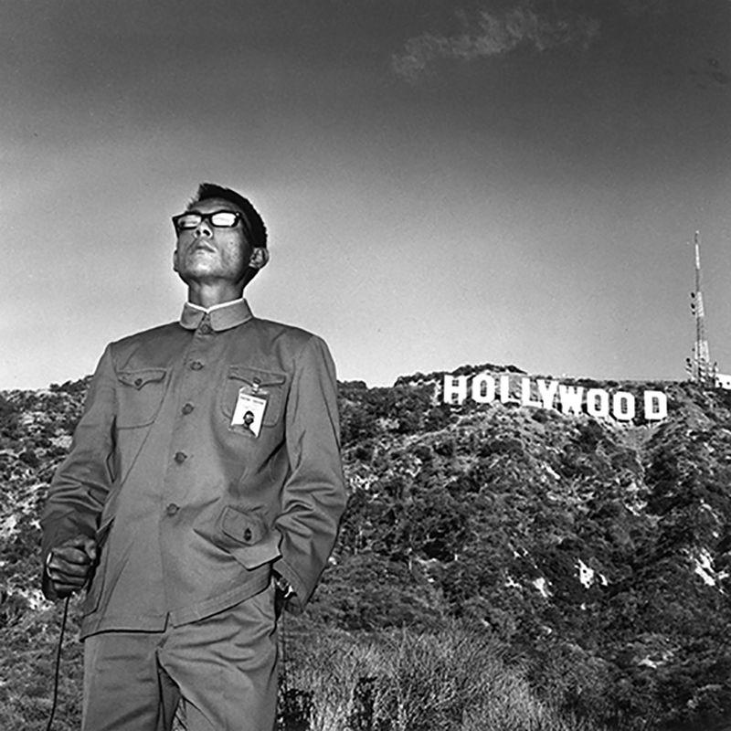 Photograph; A suited man wearing sunglasses to the left of the Hollywood sign amid the hills in the background in a black and white image.