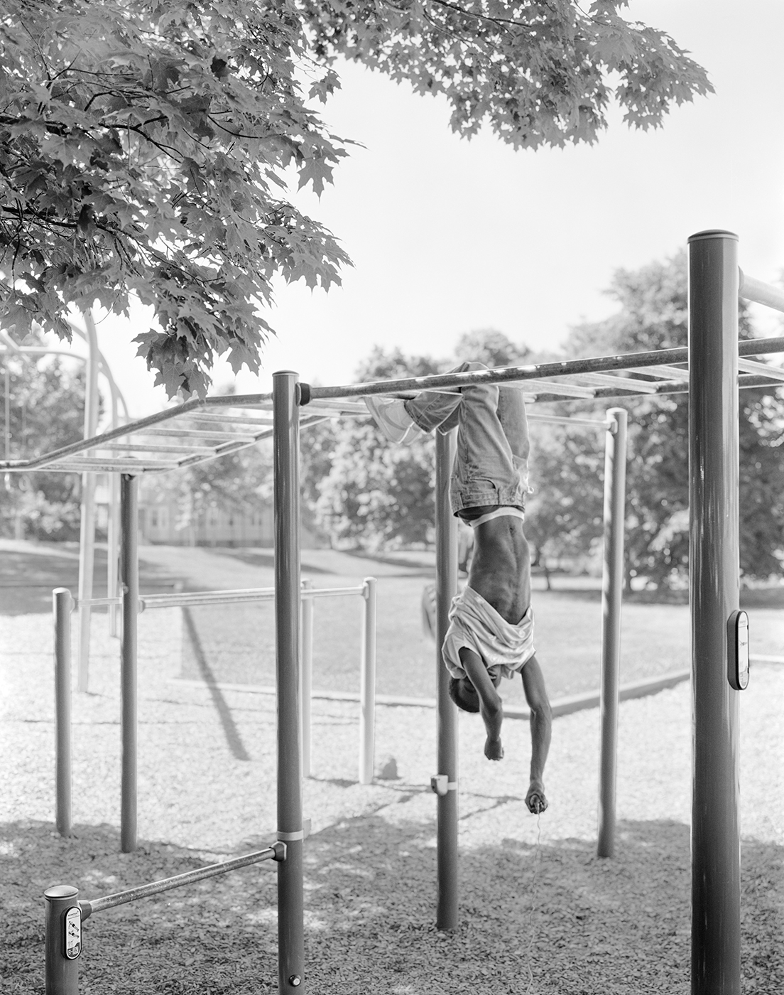 Photograph; a Black person hanging upside down from monkey bars in a playground.