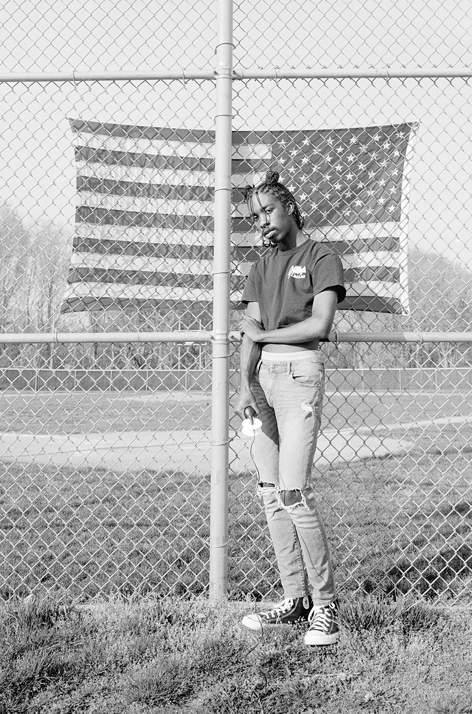 Photograph; a Black person standing in front of a chain link fence with an inverted American flag behind the fence.