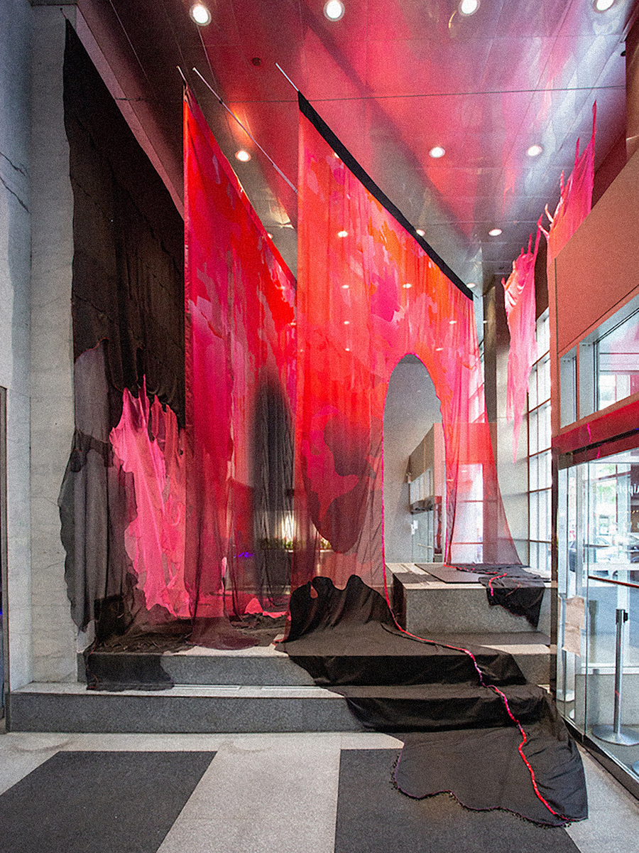 A gallery space with gray raised steps and large red and black gauzy fabric hanging down onto the steps. The fabric has some darkened silhouettes and gaps.