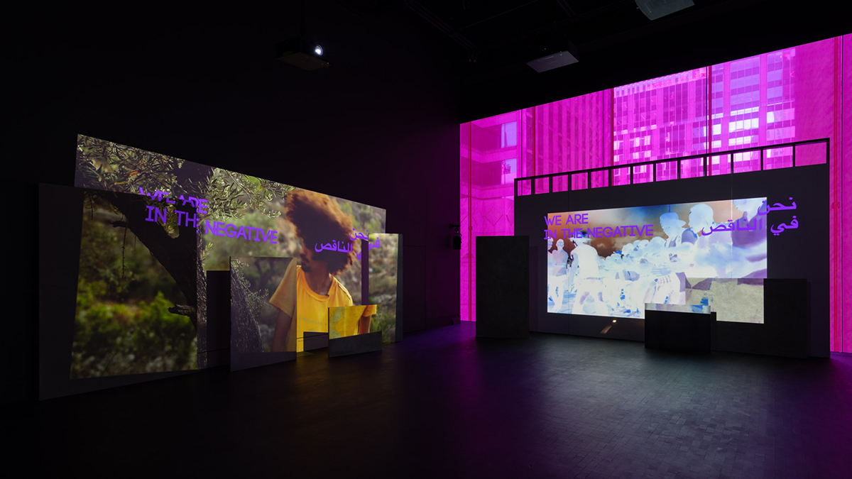 A dark gallery space with bright images projected onto screens along with purple colored text reading "we are in the negative" in English and Arabic superimposed over the images.