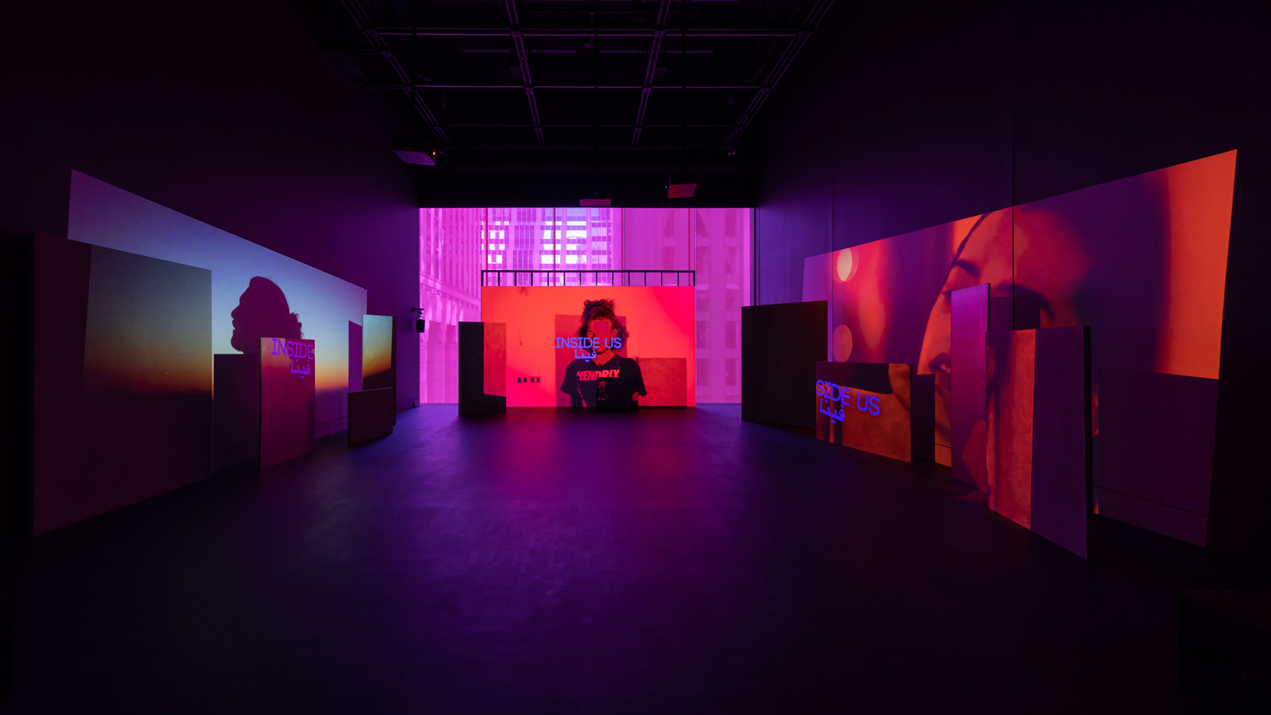 A dark gallery space with colorful images including people's faces projected onto the walls and purple text reading "Inside Us" in English and Arabic.